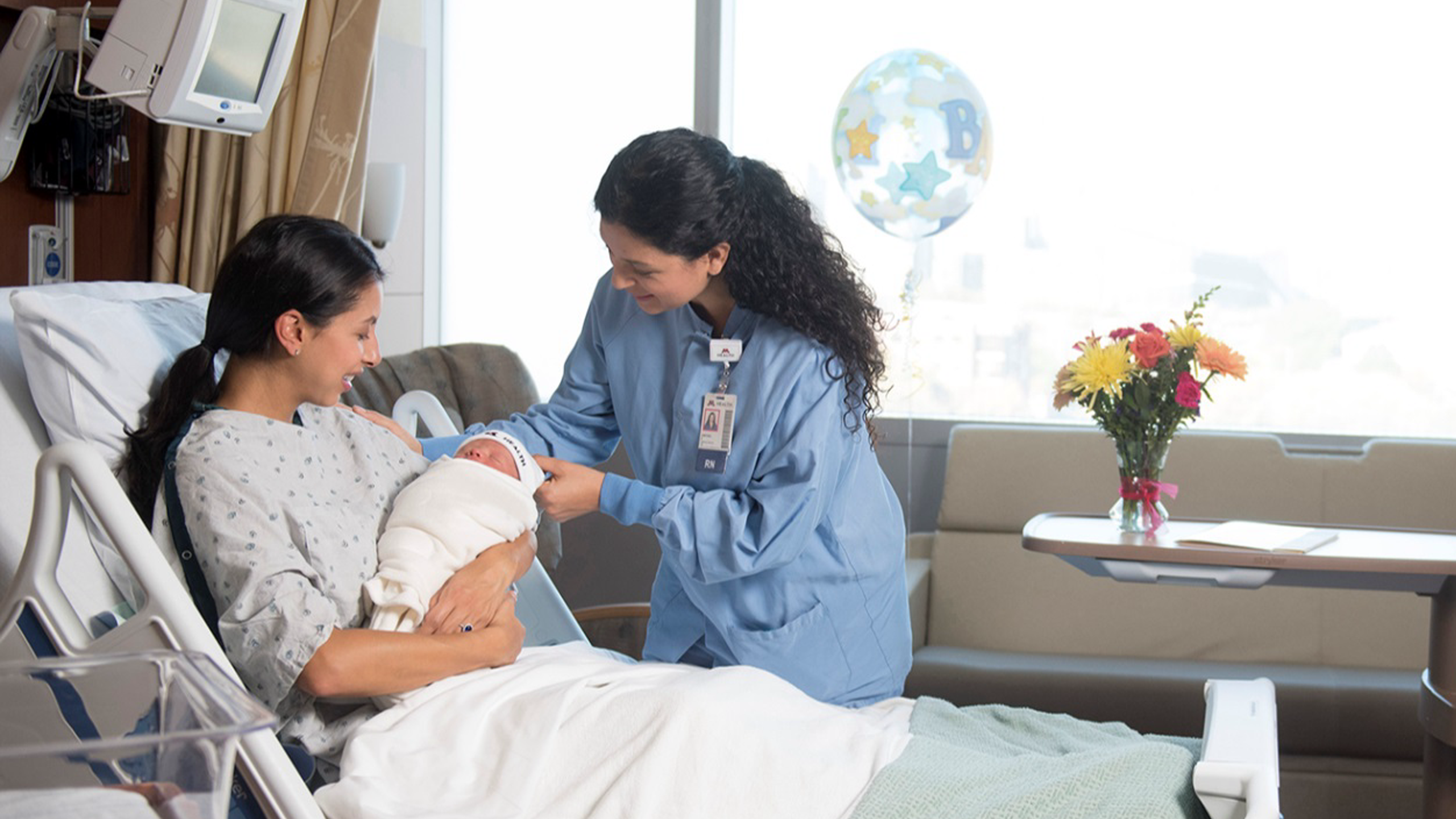 Mother in hospital holding newborn baby while a nurse looks on.